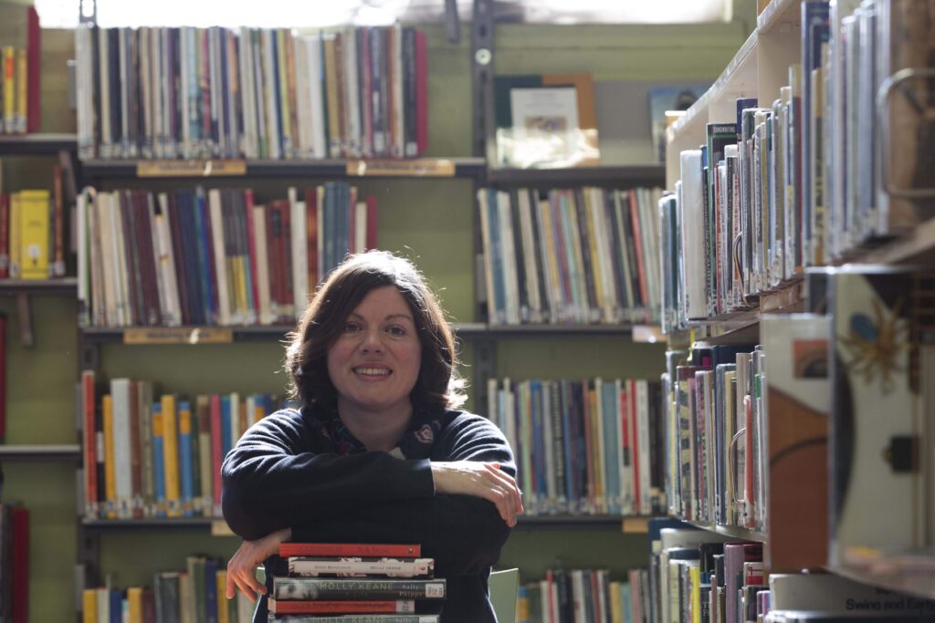 Kathy is sitting at a table resting her arms on a stack of books in the Cork City Library