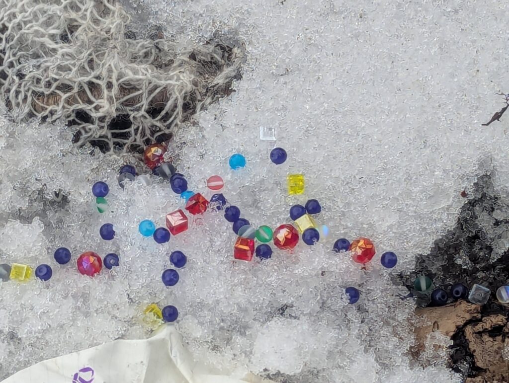 Photograph of snow on old tree roots: some off-white lacy knitting and colourful beads are scattered into the snow. Some of the beads appear to be roughly in the shape of the infinity symbol, used by many as the symbol for autism.