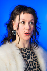 Aby is a white person with long dark wavy hair. She is wearing a leopard-print top and a white furry jacket and standing in front of a bright blue background.