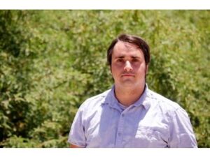 Dave is a white man wearing a pale shirt, standing in front of a group of trees in the sun.