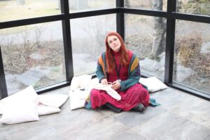 Megan is a white woman with long red hair, dressed in dark red and green. She is sitting on the ground in an empty greenhouse.