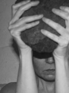 Black and white photo of a woman from the neck up, with a serious expression, holding a large rock in front of her face.