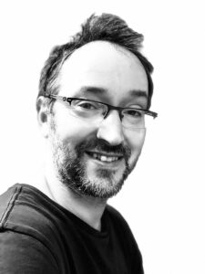 William is a white man with very short dark hair and a short beard. He is wearing glasses and a dark short and is smiling.