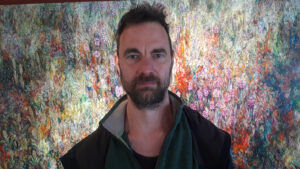 Ole is a white man with short dark hair and a short beard. He is wearing a dark fleece top and standing in front of a brightly lit, colourful background.